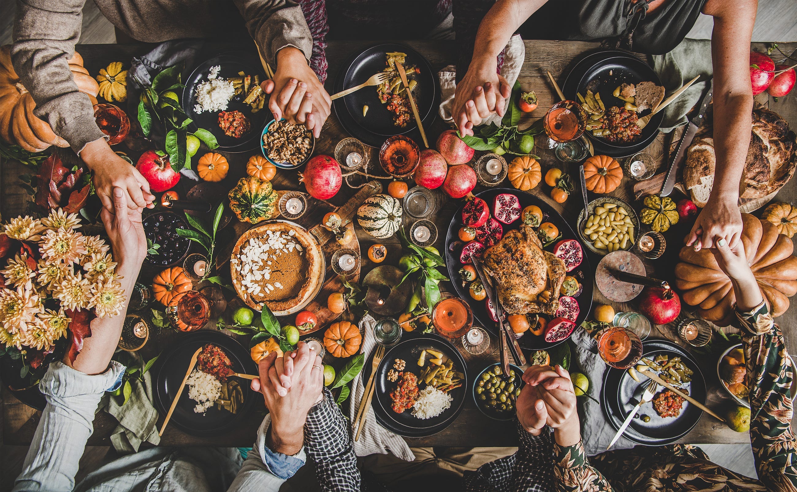 Family praying holding hands at Thanksgiving table. Flat-lay of feasting peoples hands over Friendsgiving table with Autumn food, candles, roasted turkey and pumpkin pie over wooden table, top view -By sonyakamoz Family praying holding hands at Thanksgiving table. Flat-lay of feasting peoples hands over Friendsgiving table with Autumn food, candles, roasted turkey and pumpkin pie over wooden table, top view -By sonyakamoz