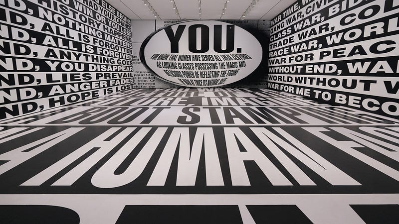 The view is from the center of a rear wall, looking at the surrounding walls and floor, which stretch far ahead. All of them are covered in massive, black-and-white text. The wall opposite has giant, slightly curved text encapsulated within an oval: “YOU. You know that women have served all these centuries as looking glasses possessing the magic and delicious power of reflecting the figure of man at twice its natural size.” The view is from the center of a rear wall, looking at the surrounding walls and floor, which stretch far ahead. All of them are covered in massive, black-and-white text. The wall opposite has giant, slightly curved text encapsulated within an oval: “YOU. You know that women have served all these centuries as looking glasses possessing the magic and delicious power of reflecting the figure of man at twice its natural size.”