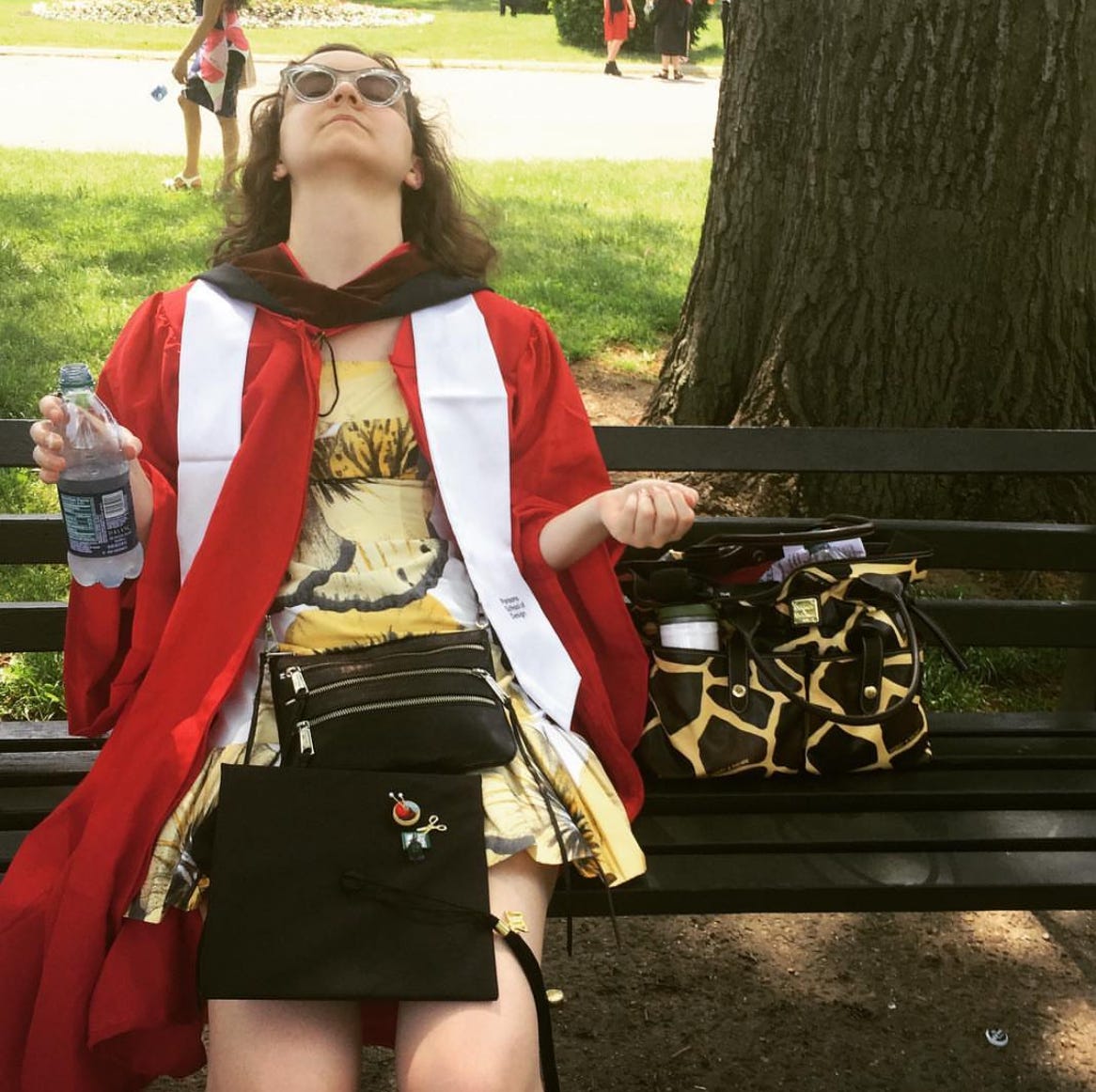 Young woman in a yellow, floral dress looking exhausted on a bench. She has graduation regalia from Parsons on - a red cap and a black gown.