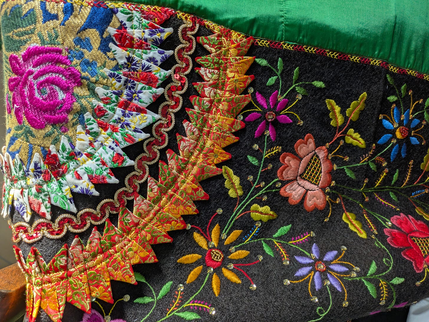 Close-up of a richly embroidered textile featuring vibrant floral designs and geometric patterns in bright pink, gold, green, and multicolor threads on a dark fabric background.