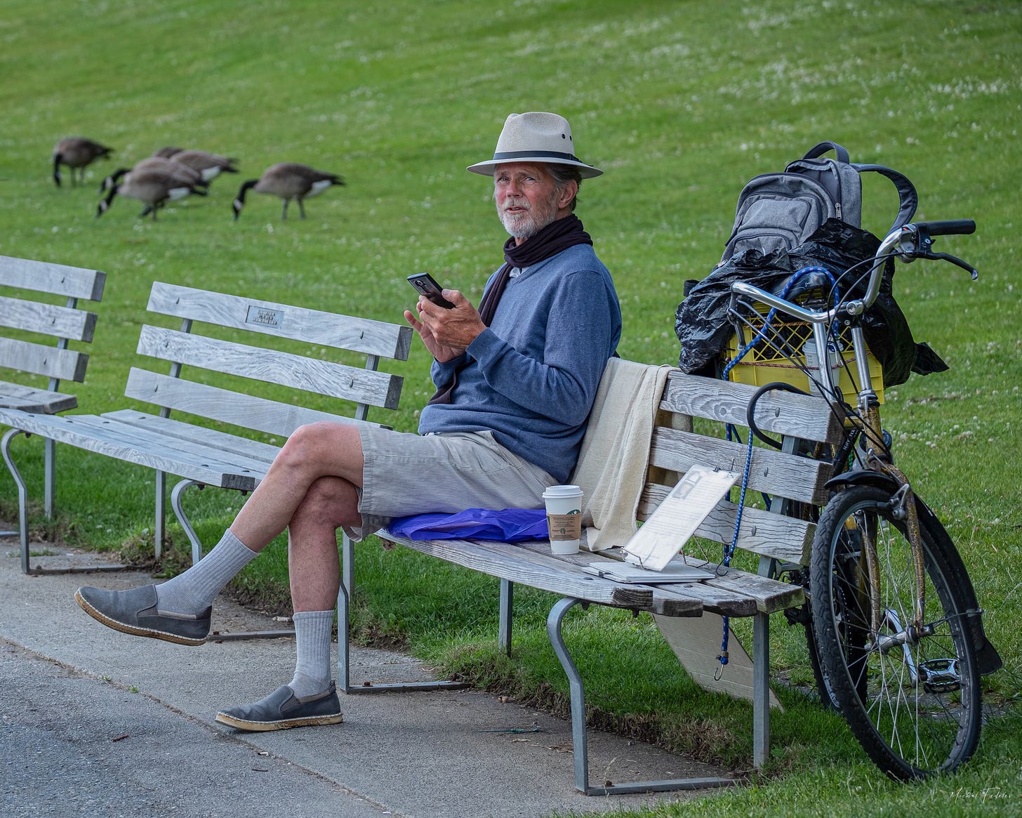 A man in shorts and a hat sitting on a park bench, with a bicycle leaning against the bench and Canada geese in the background.