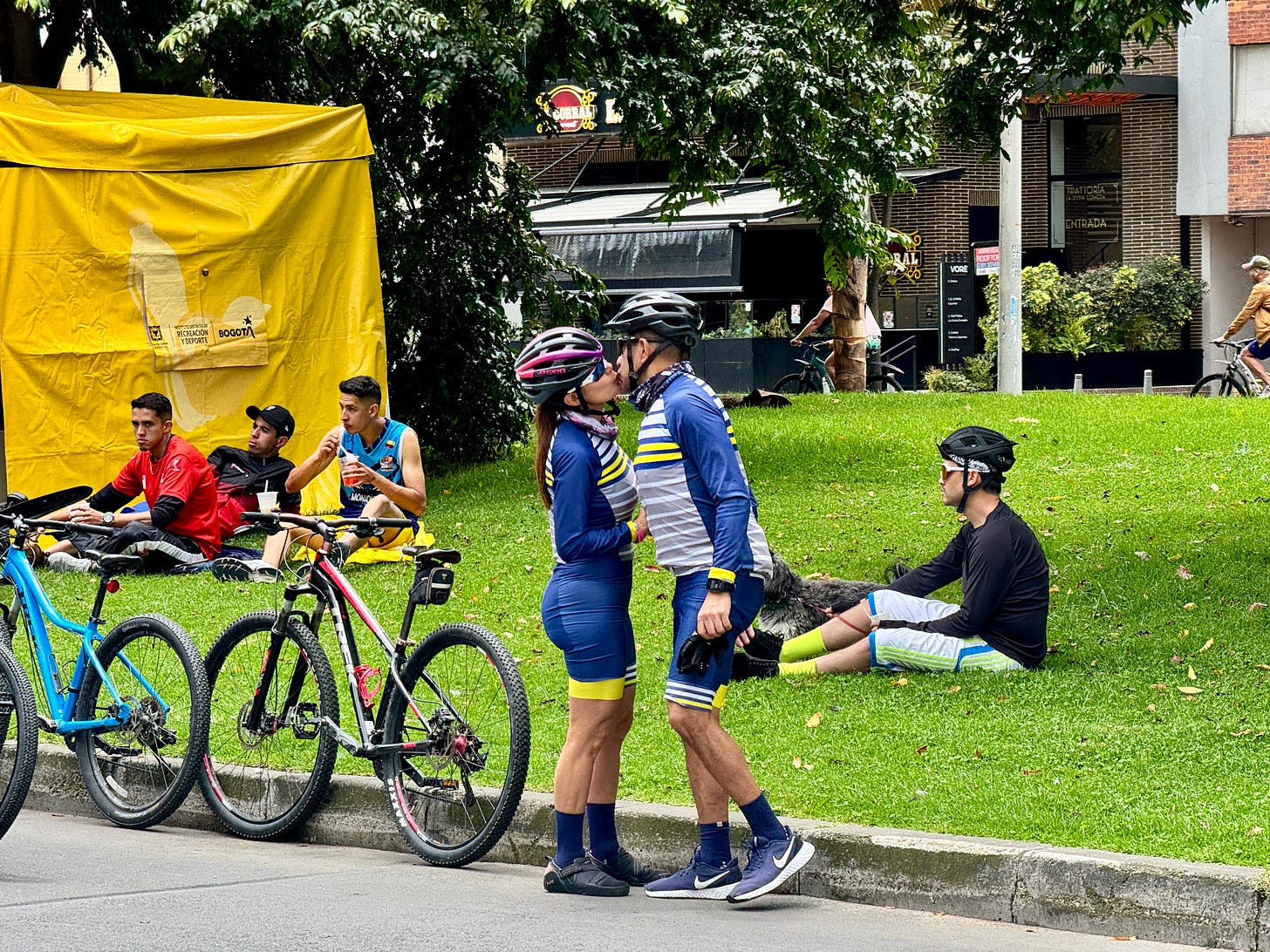 Dos personas besándose vestidos de ciclistas mientras otros descansan sobre el césped. Dos personas besándose vestidos de ciclistas mientras otros descansan sobre el césped.