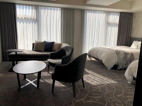 A modern and spacious hotel room with twin beds, a seating area with armchairs and a chaise lounge, a flat-screen TV, and large windows with sheer curtains. The accessible bathroom features grab bars, a handheld showerhead, and a shower chair.