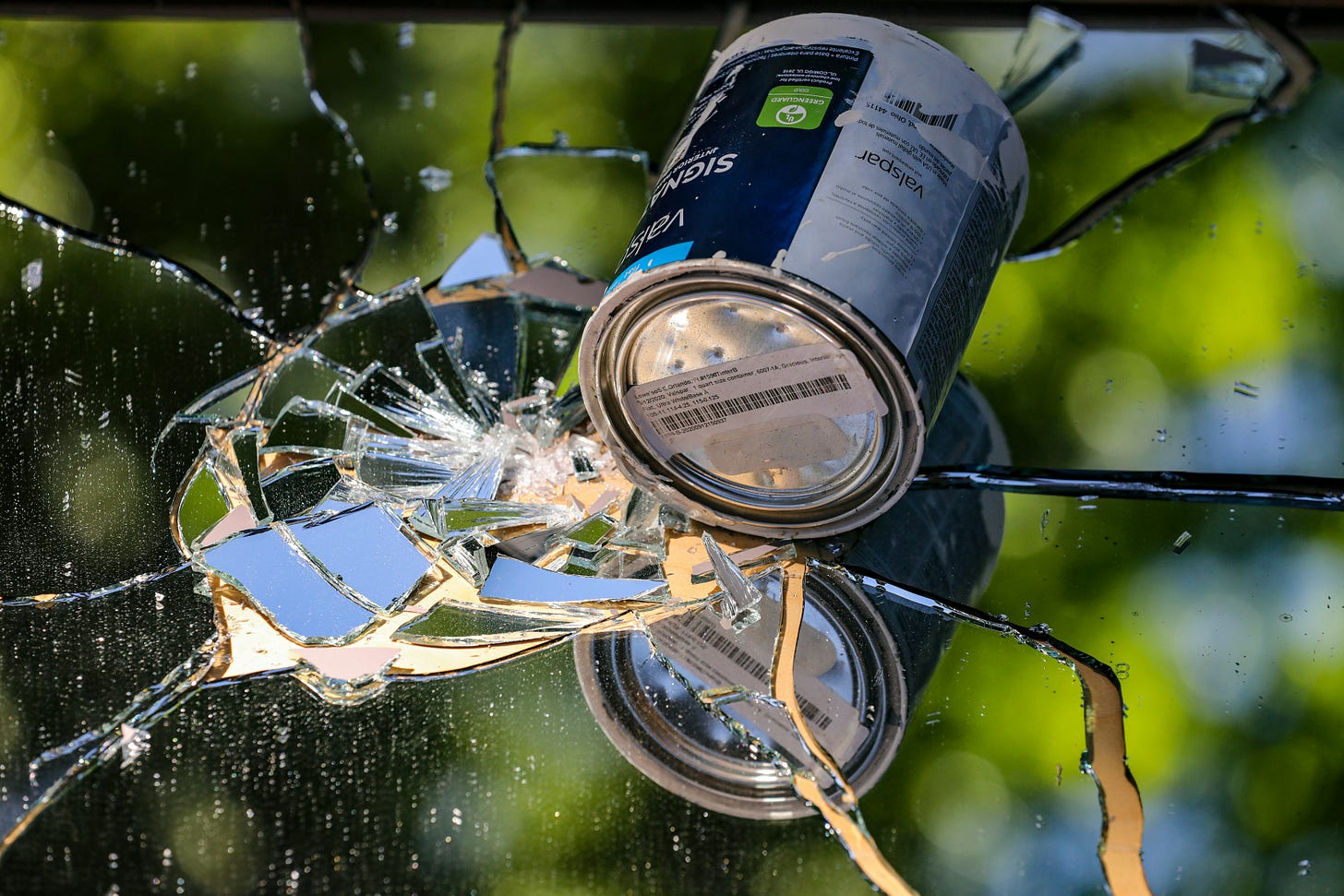 A can of paint smashes into a mirrored pane of glass. In the background are light and dark green shapes suggesting a tree/sky scene.