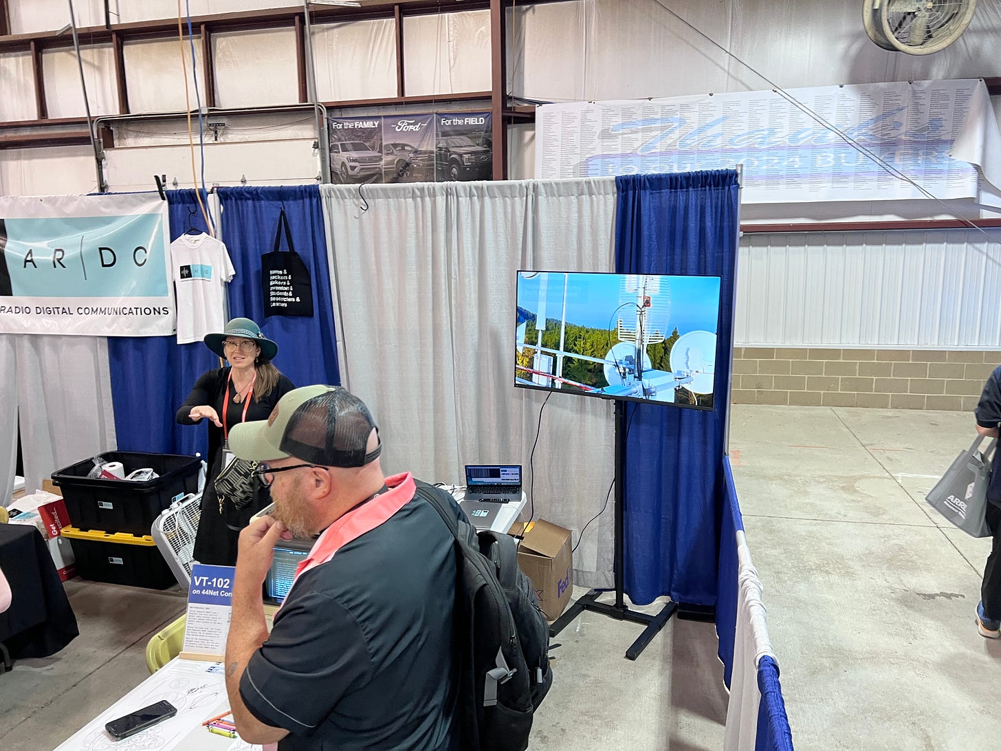 Rosy Schechter KJ7RYV at Hamvention 2026 at the ARDC booth, with (unknown) in foreground. KJ7RYV really rocks the hats! Image courtesy of Steve Stroh N8GNJ.