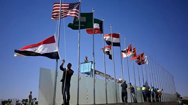 عمال يرفعون أعلام دول يزور قادتها شرم الشيخ. الصورة التقطت نهاراً، السماء صافية.