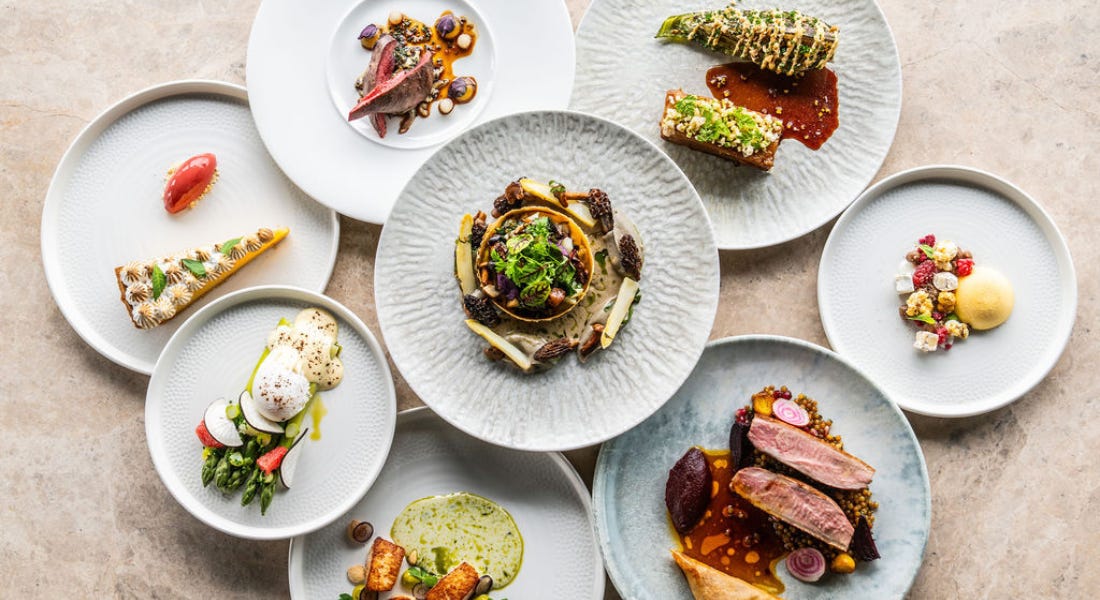 A selection of white and grey plates with colourful food plated onto them, on top of a grey stone worktop
