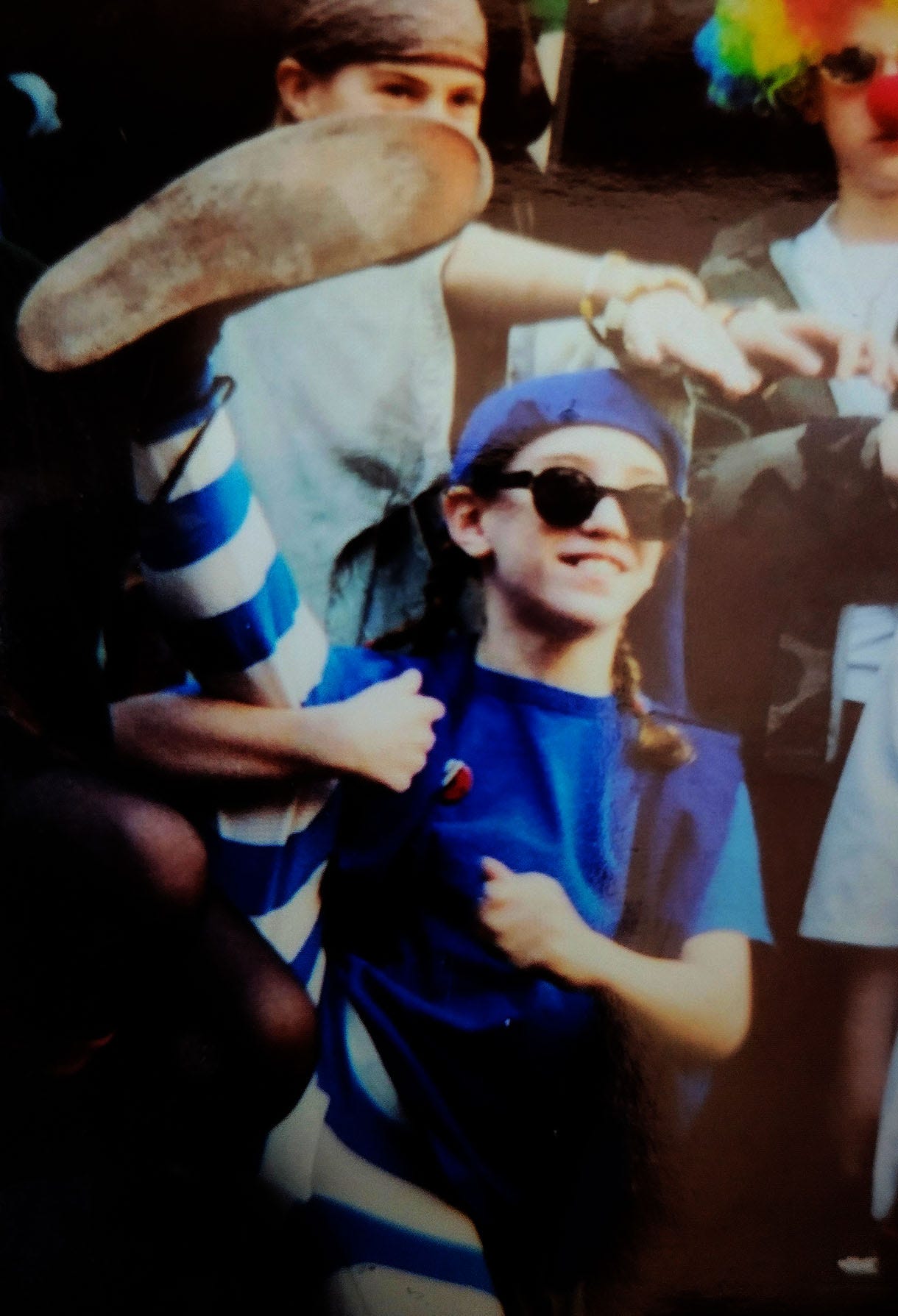 10-year old Lara in sunglasses and baggy blue and white-striped tights holds up a leg with an oversize clown shoe at the end. 