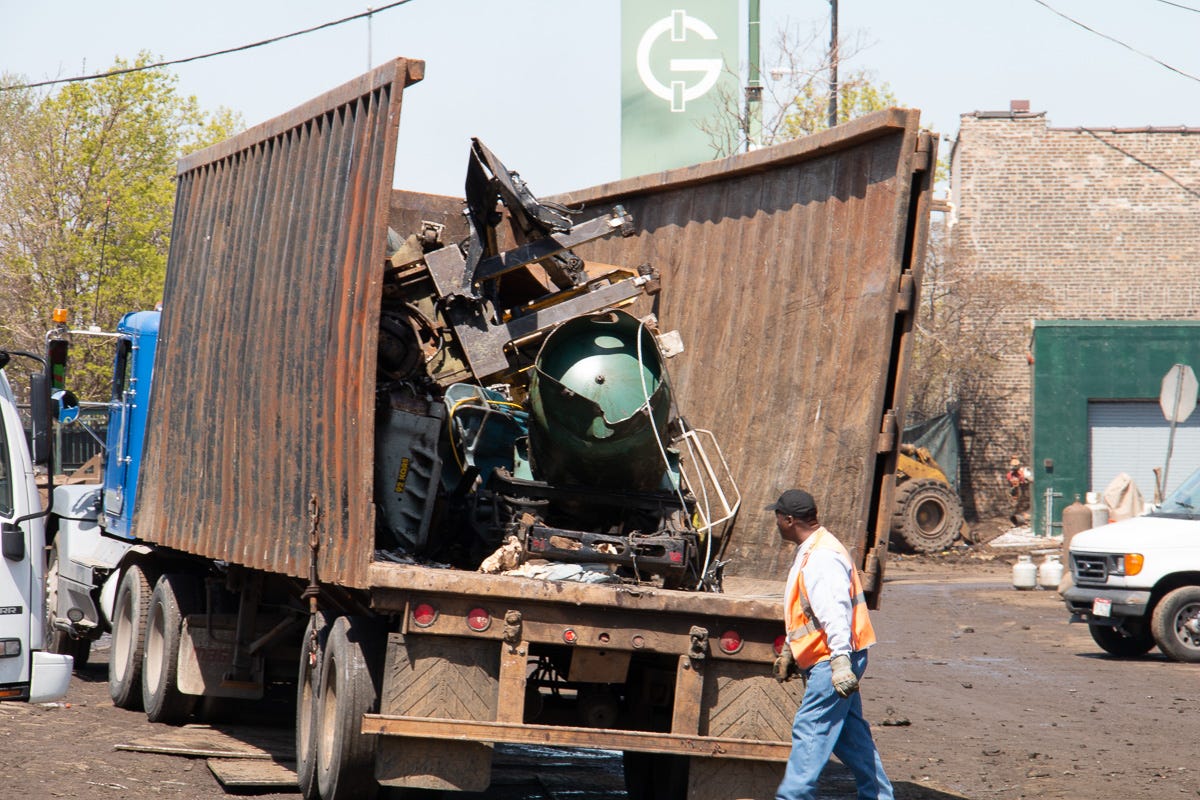 A dump trailer tilted up, offloading bulky industrial scrap while a worker in a vest walks by.