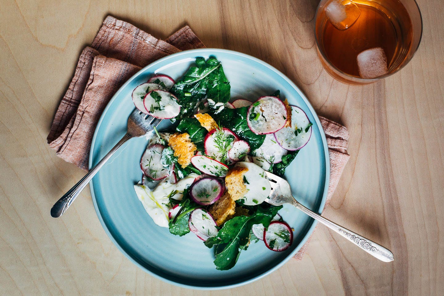 A small plate with a serving of salad and two forks. There’s a drink and a napkin alongside. 