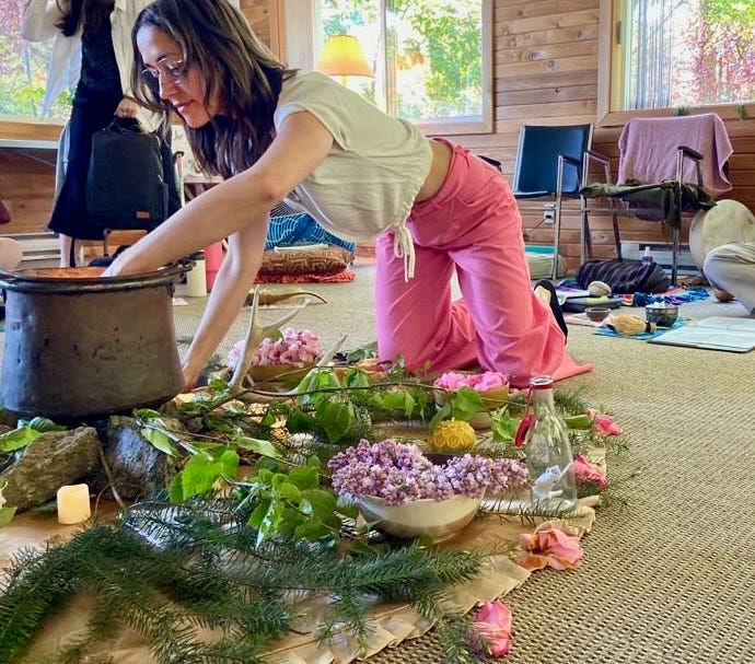 Rayann is wearing pink pants and white shirt, kneeling on the floor reaching into a cauldron, it glows from within. the cauldron is on rocks on a round cloth covered in birch, led tea lights, pink petals and other offerings