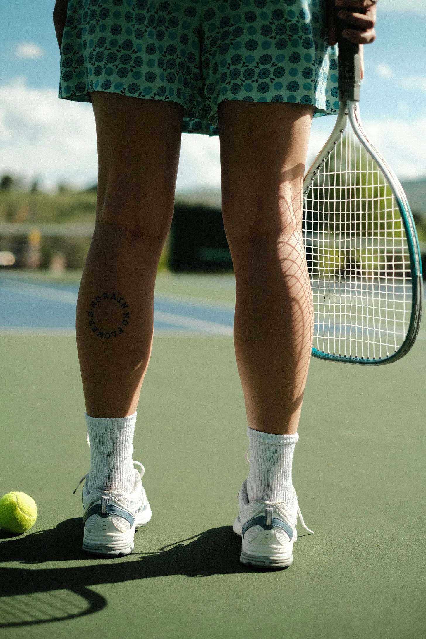 Back of a woman's legs in green shorts holding a green tennis racket.