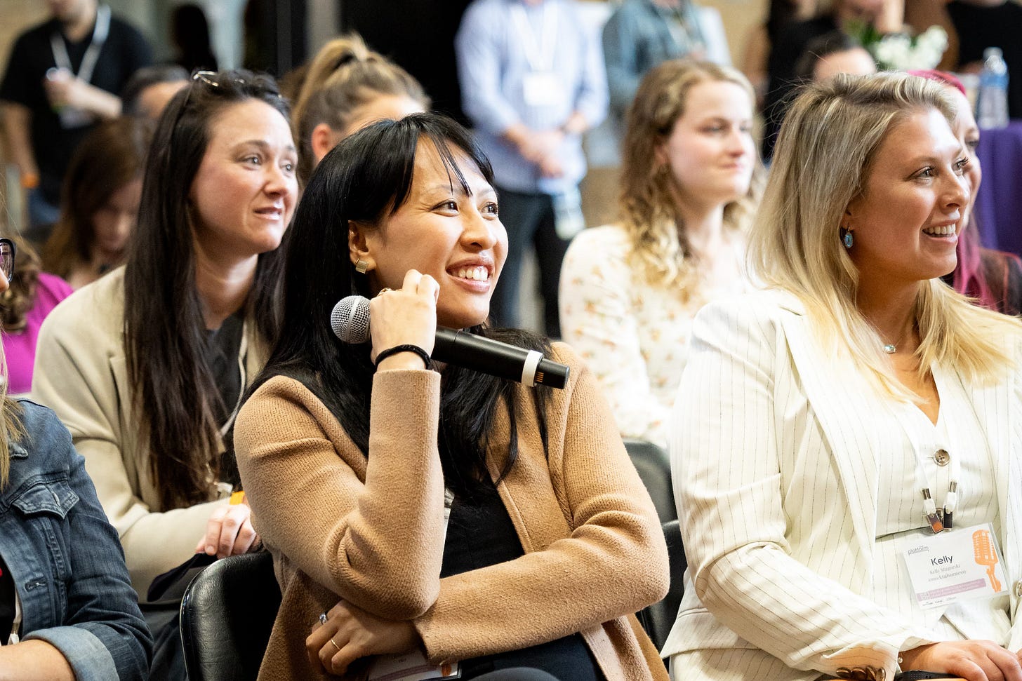 A woman smiles while holding a microphone during a panel or Q&A session at a professional event, seated among an engaged audience of women. Everyone appears attentive, relaxed, and enjoying the conversation.