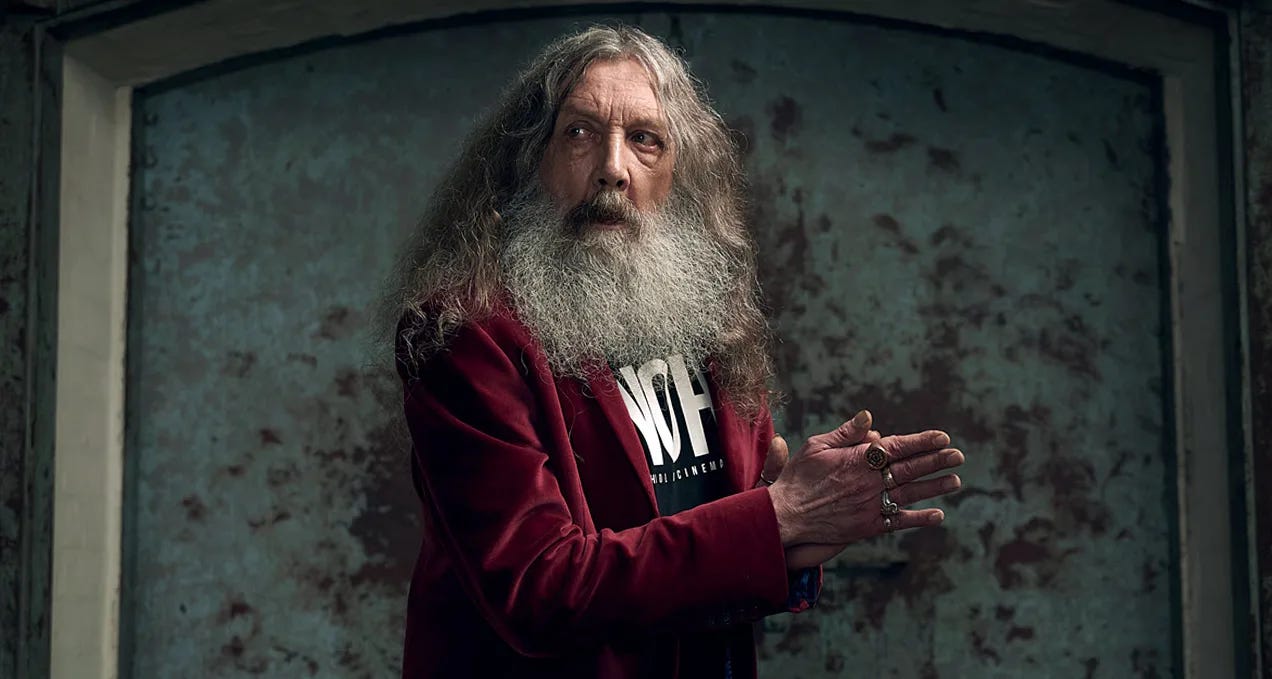 Um homem idoso, com longo e farto cabelo grisalha, barba ainda mais farta e também grisalha, camiseta preta com algum texto branco, um blazer cor de vinho e as mãos cheias de anéis. Ele está meio na diagonal e olhando para o lado. Um homem idoso, com longo e farto cabelo grisalha, barba ainda mais farta e também grisalha, camiseta preta com algum texto branco, um blazer cor de vinho e as mãos cheias de anéis. Ele está meio na diagonal e olhando para o lado.