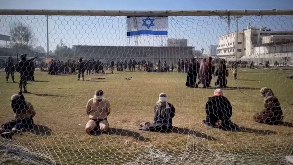 A still from a video appears to show Palestinian women and men blindfolded with their hands tied behind their backs as they sit on the grass in front of a soccer goal in the stadium. A still from a video appears to show Palestinian women and men blindfolded with their hands tied behind their backs as they sit on the grass in front of a soccer goal in the stadium.