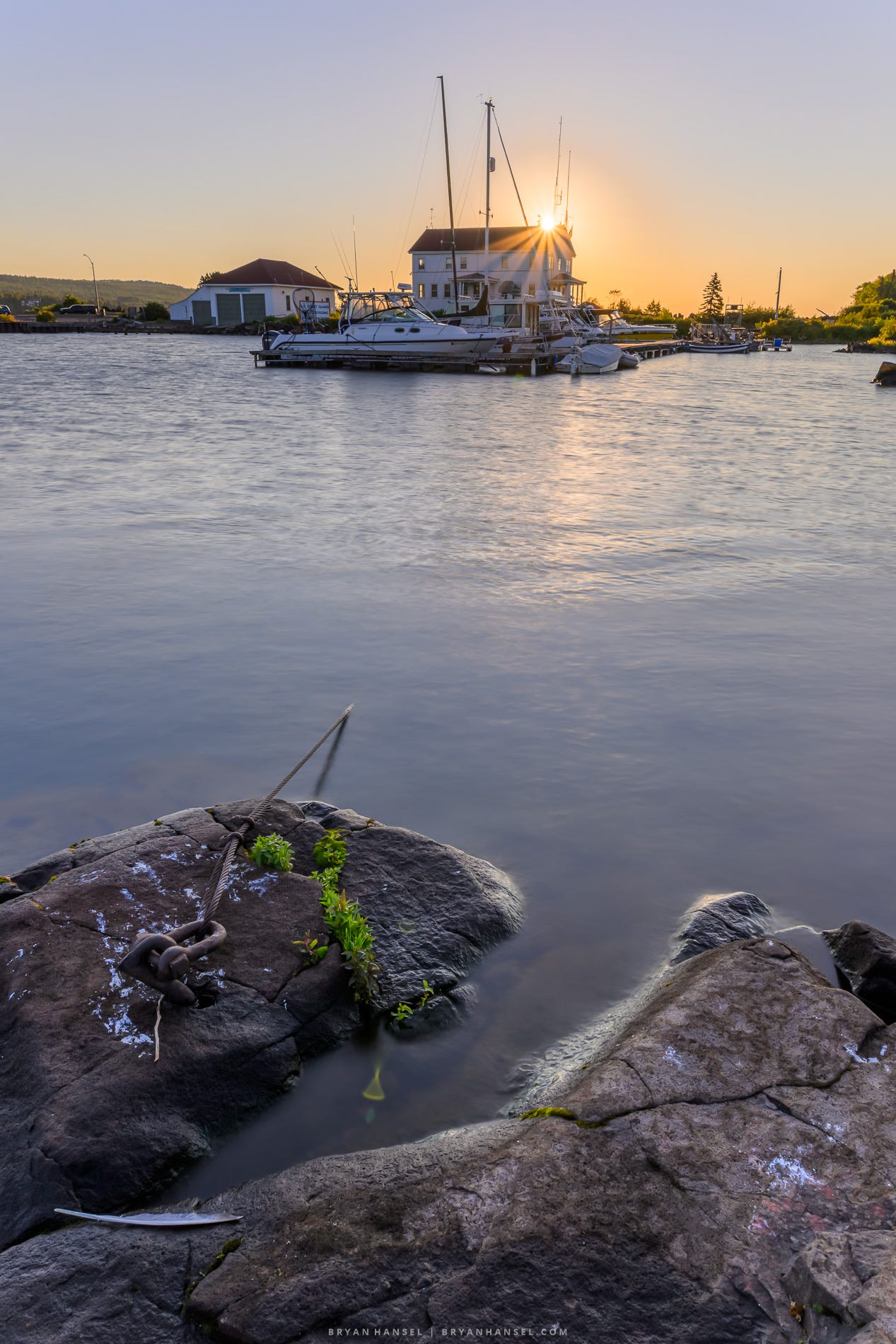 Sunrise over the Grand Marais harbor, with boats moored in front of the Coast Guard building. Smooth water reflects the warm morning light, while granite shoreline rocks and a mooring chain fill the foreground. HDR and auto