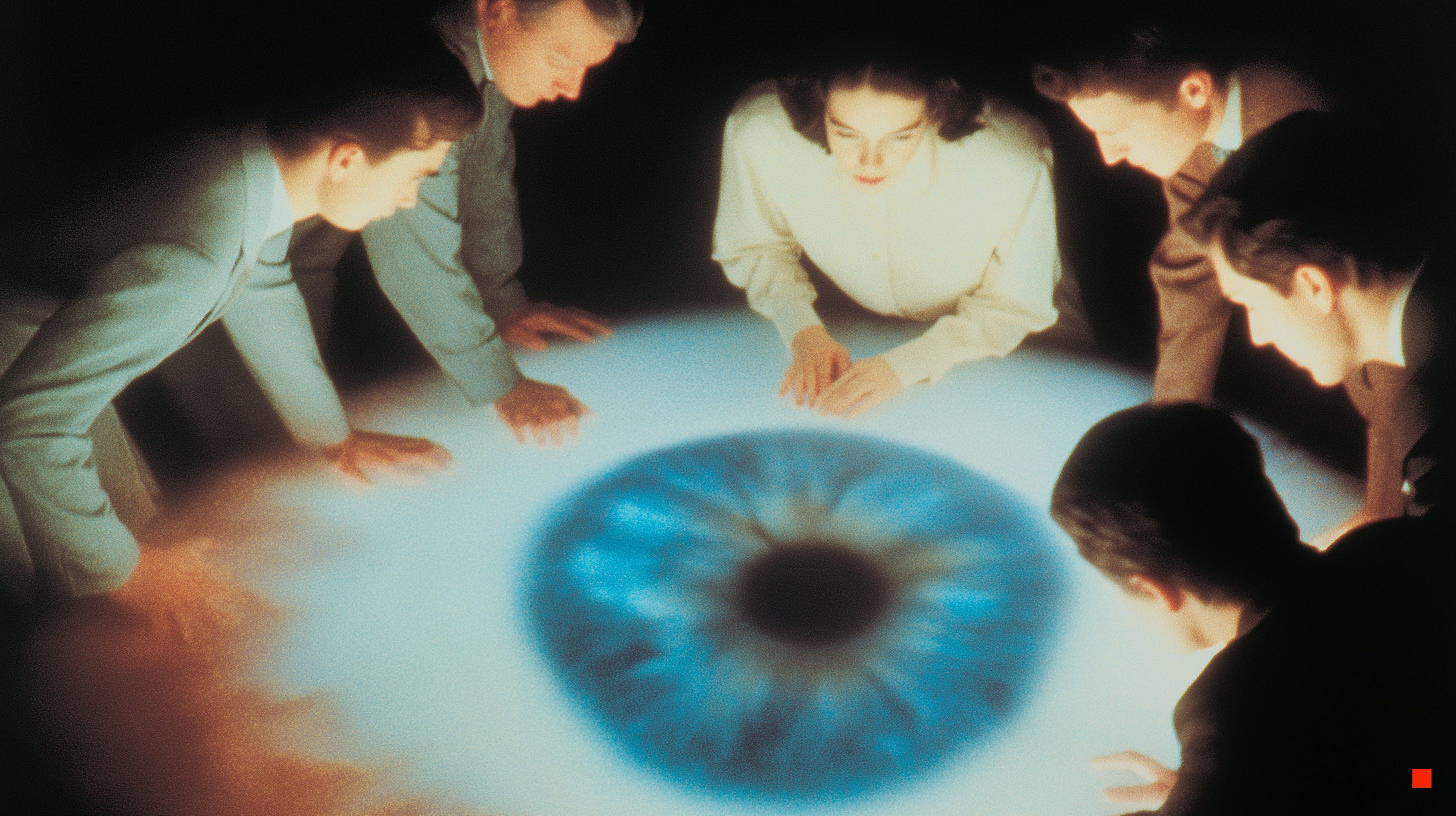 A group of people gathered closely around a large projected human eye, viewed from above, each person leaning inward toward the image in a darkened space.