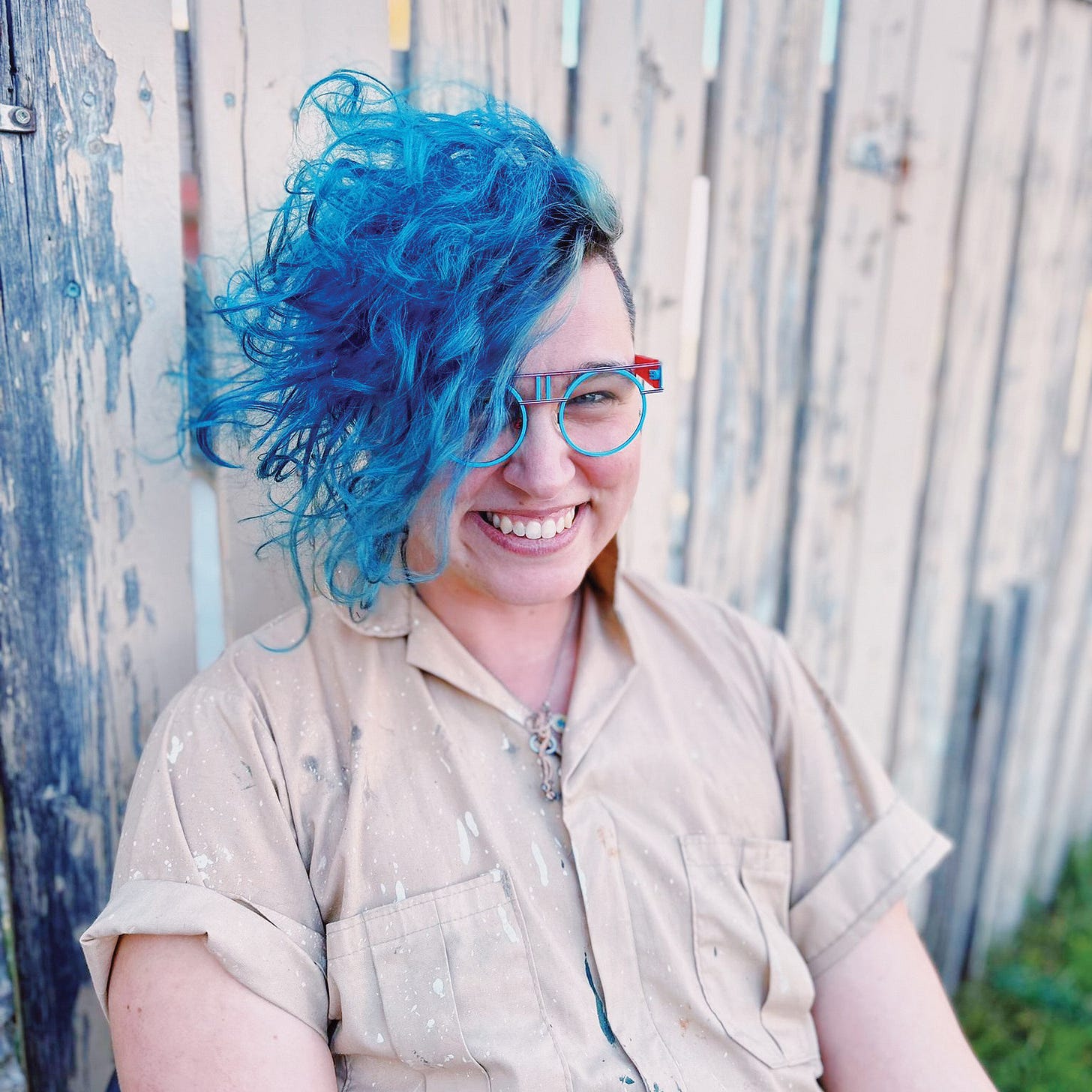 A person with blue hairs sits by a fence and smiles at the camera A person with blue hairs sits by a fence and smiles at the camera