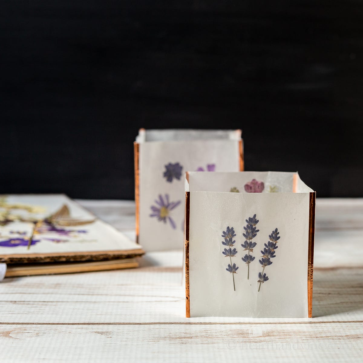 A photo of small white square envelopes with shiny rose gold edges are decorated with pressed flowers in pastel purples and pinks in a dark black studio with white wooden table.