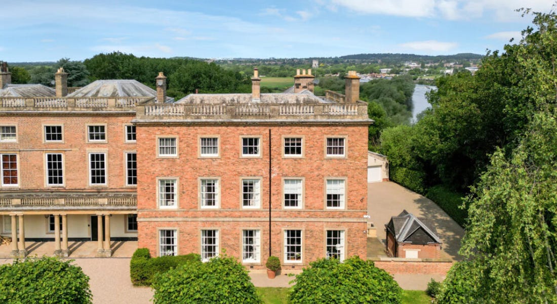 A large brick stately home with greenery around it and a blue sky above it