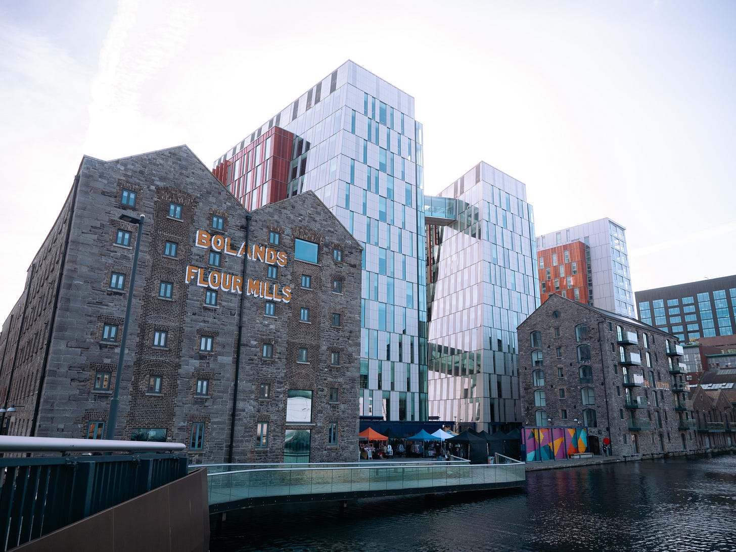 A view from the dock: the old brick factory facade of the mills is adorned with yellow lettering that reads Bolands Flour Mills. It overlooks the water, while large glass buildings loom behind housing the Google offices. 