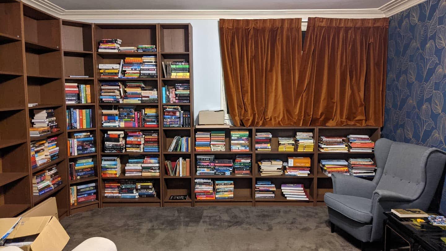 A room with a curtained window, the walls covered in floor to ceiling bookshelves; books cover most of the shelves, and there's an armchair to one side.