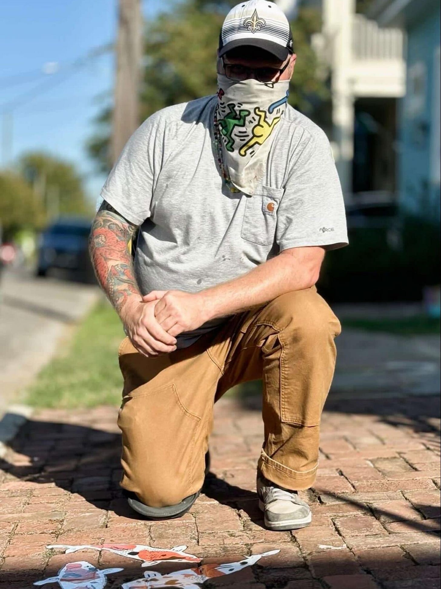 Jeremy is a white man wearing a baseball cap and bandana covering his nose and mouth with Keith Haring artwork on it. He is kneeling on a brick sidewalk, shadowed in sunlight. He wears a gray T-shirt and brown pants and grey shoes, with a tattooed right arm. He is posing with his koi fish artwork on the bricks in front of him. He is holding his hands together, with his left arm resting on his left knee. There are houses and a street blurred in the background behind him. Jeremy is a white man wearing a baseball cap and bandana covering his nose and mouth with Keith Haring artwork on it. He is kneeling on a brick sidewalk, shadowed in sunlight. He wears a gray T-shirt and brown pants and grey shoes, with a tattooed right arm. He is posing with his koi fish artwork on the bricks in front of him. He is holding his hands together, with his left arm resting on his left knee. There are houses and a street blurred in the background behind him.