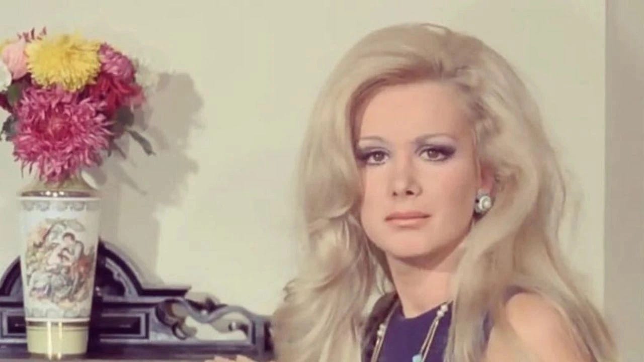 Filiz Akın, arka planda çiçekli bir vazo ile şık bir iç mekanda, zarafetiyle Yeşilçam'ın unutulmaz yıldızlarından biri.