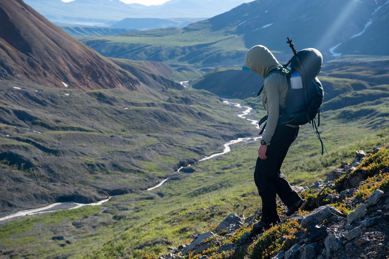 A hooded hiker leans forward against the weight of a pack on a steep tundra shoulder, gazing down into a deep green valley where a braided meltwater stream winds between rounded, grass-covered hills. Low sun rakes across the slopes, and distant ridges fade into haze at the head of the valley.