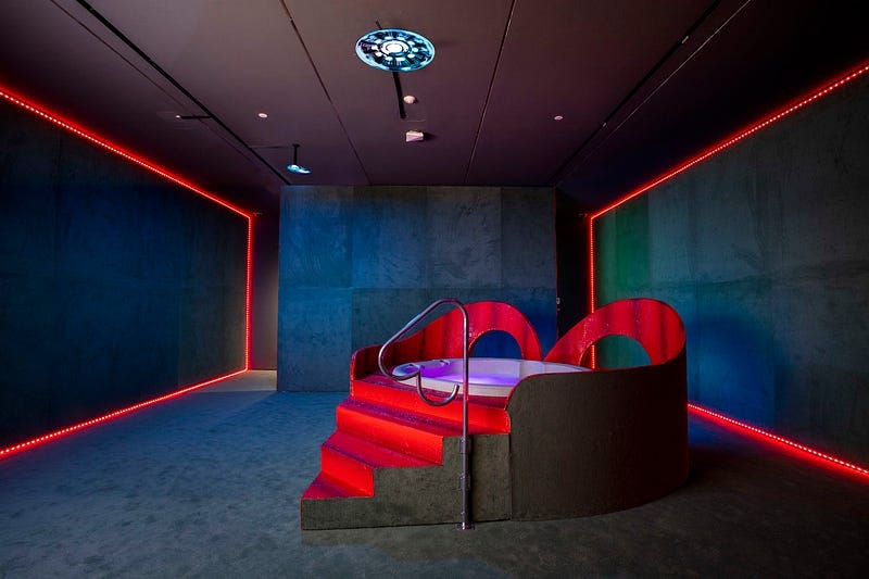 A dim room with dark grey carpeting and concrete walls. Thin tracks of red lighting line the edges of the floor and ceiling. In the room’s center is a large, red, heart-shaped hot tub. The water has purple lighting and the red stairway has a heart-shaped steel railing to one side.