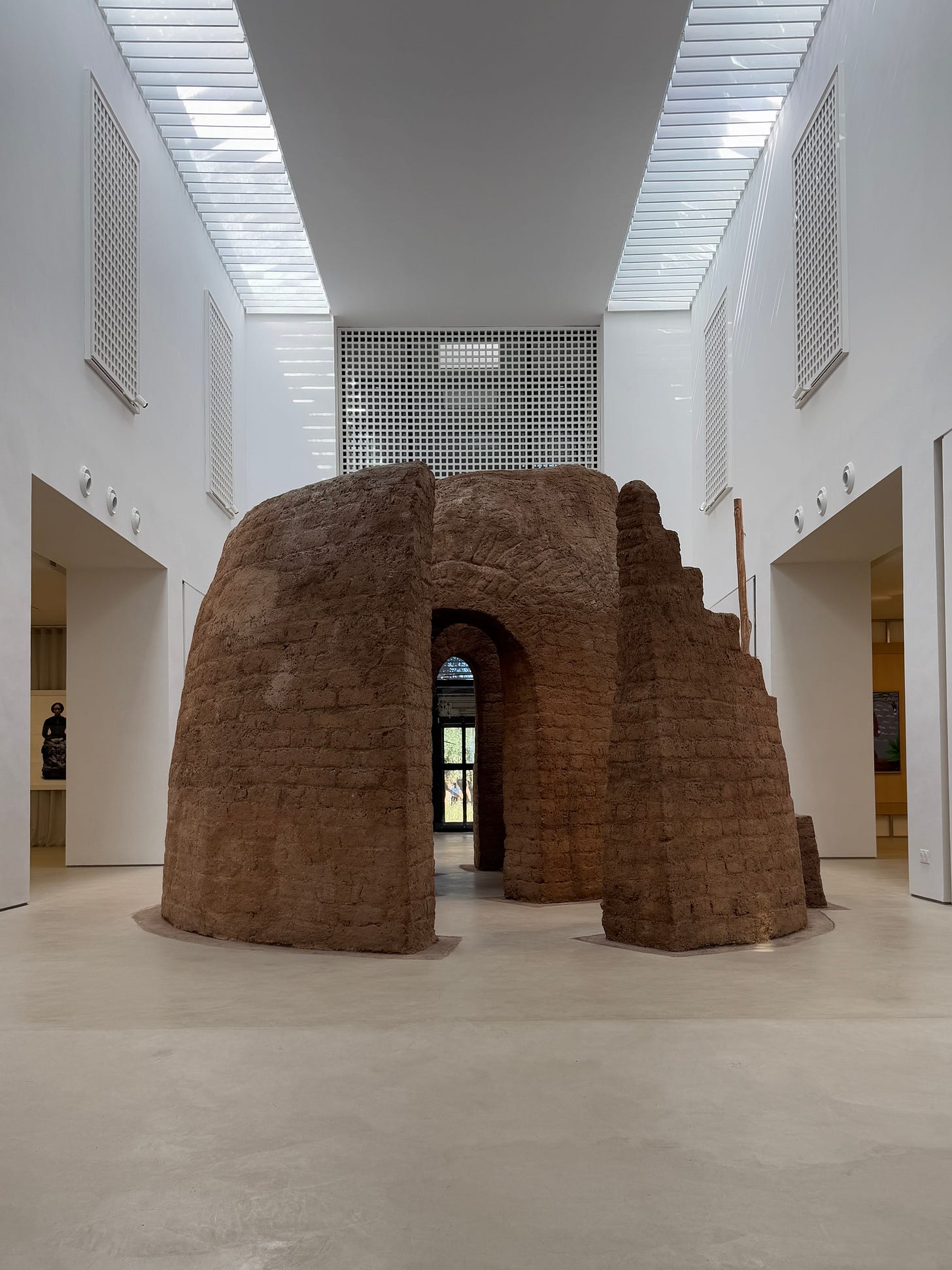 Installation Dans les bras de la terre by Salima Naji at MACAAL, Marrakech: a walk-in, dome-like structure built from earthen materials stands at the center of a bright, contemporary museum space with skylights, contrasting traditional earth-based construction with the museum’s clean, modern architecture. Installation Dans les bras de la terre by Salima Naji at MACAAL, Marrakech: a walk-in, dome-like structure built from earthen materials stands at the center of a bright, contemporary museum space with skylights, contrasting traditional earth-based construction with the museum’s clean, modern architecture.