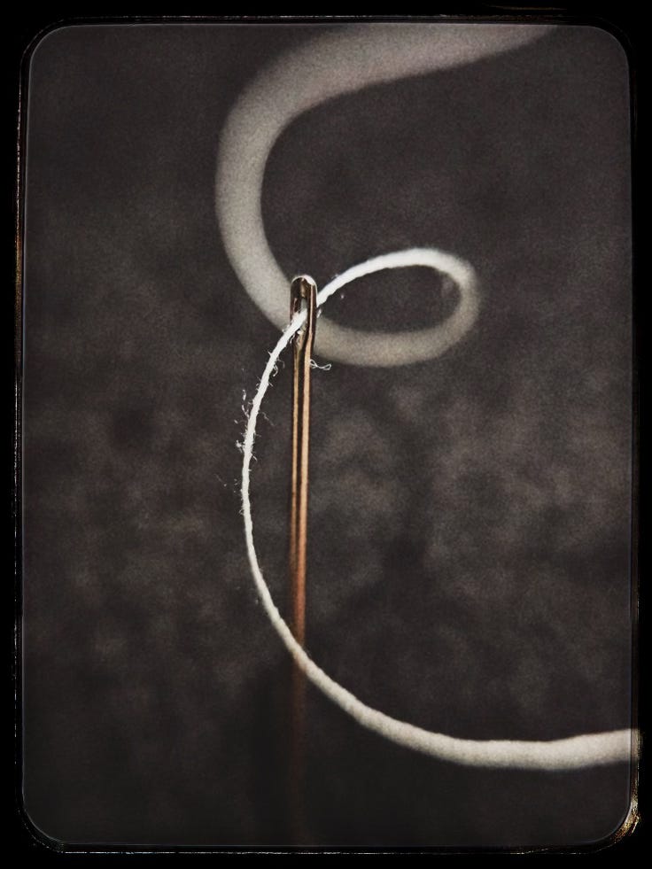 A close-up photograph of a sewing needle standing upright in dark fabric, with a pale thread looping loosely through its eye and curving softly into the background.