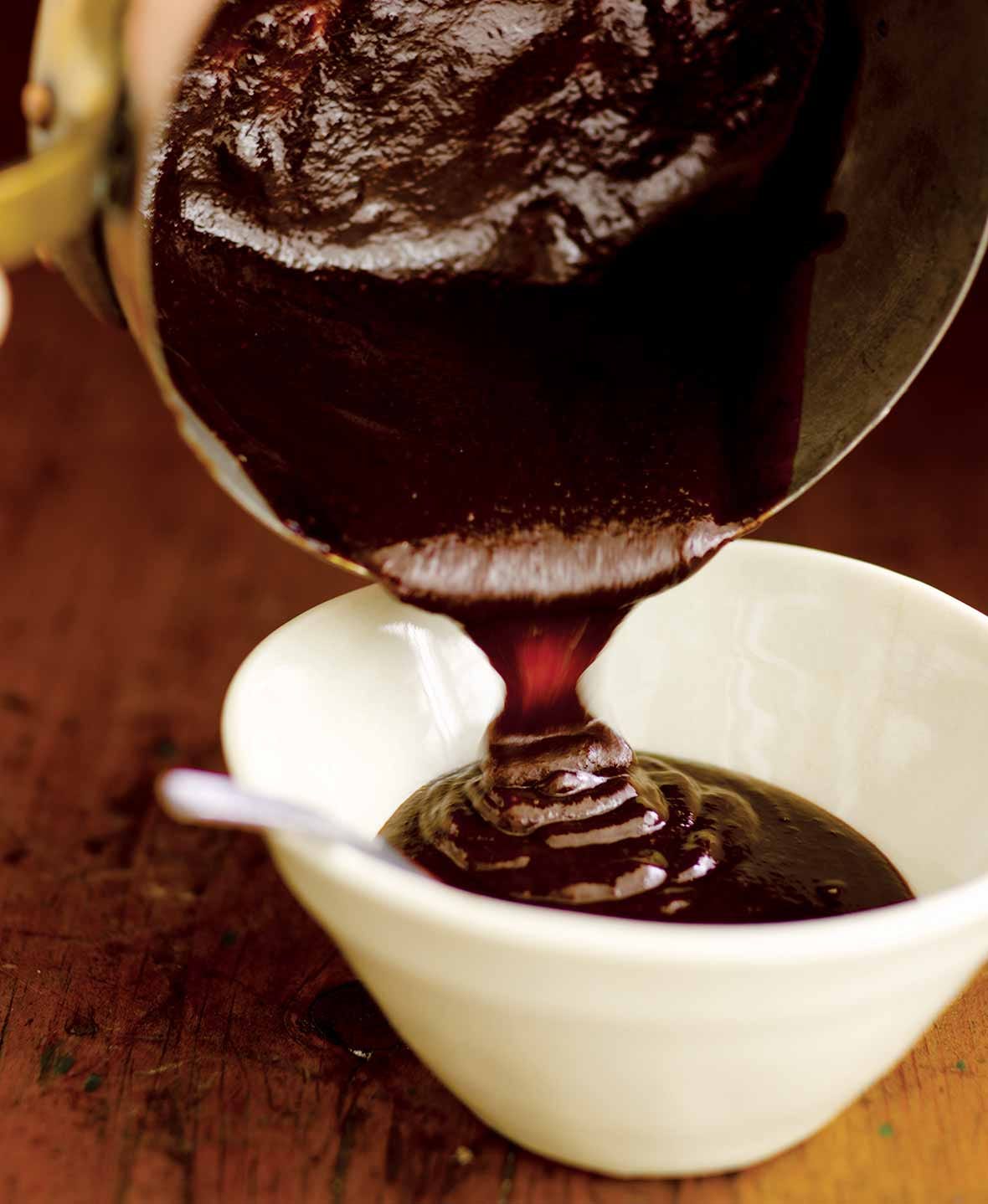 Thick, dark barbecue sauce pouring from a copper pot into a white ceramic bowl with a spoon.