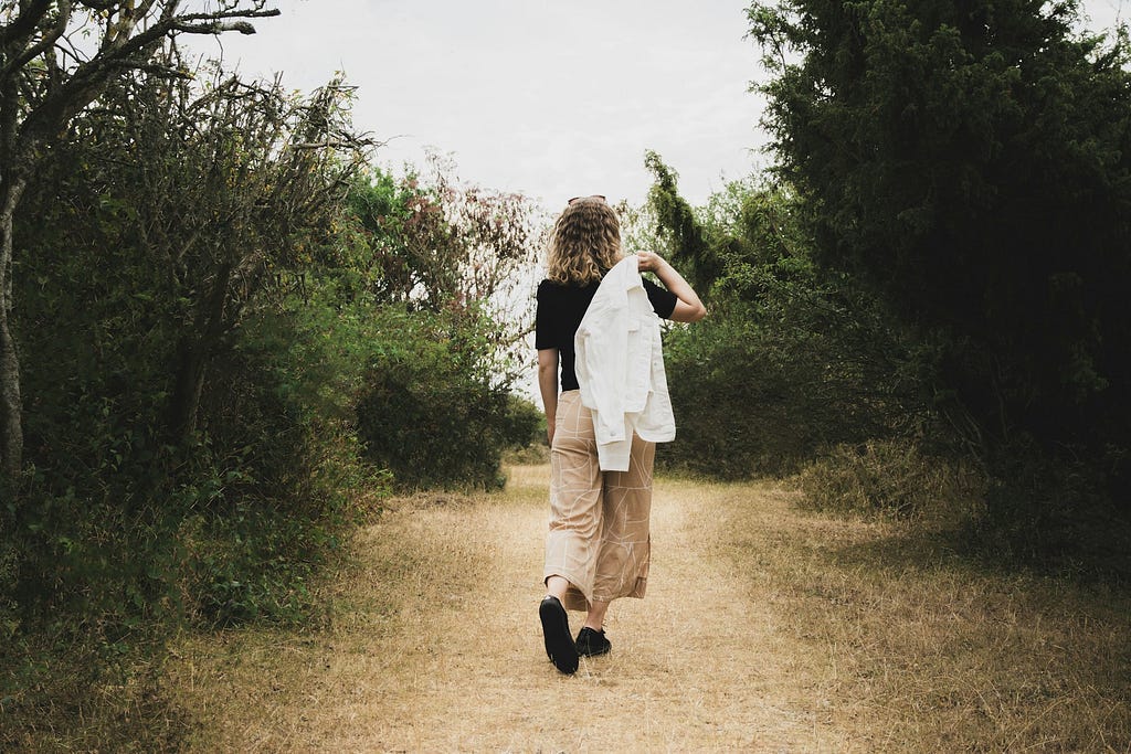 Back of a woman carrying a white dress shirt over her shoulder and walking down a dirt path surrounded by trees.