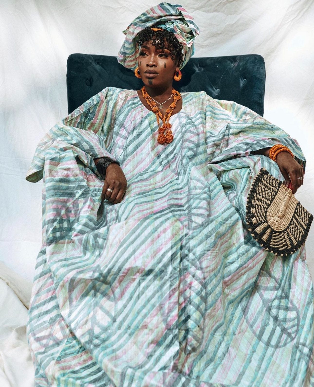 Lovette Jallow dressed in blue traditional attire, holding a ceremonial fan. Her stance reflects Fulani matriarchal elegance and the authority carried through gesture, presence, and ancestral memory.