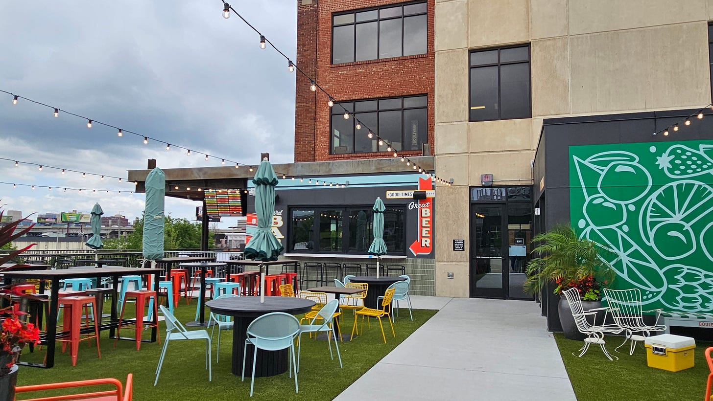 Finished Boulevard Yard deck with artificial turf, mixed orange, teal, and yellow café seating under string lights, a "Good Times Great Beer" sign, and a green cocktail mural along the building wall. Finished Boulevard Yard deck with artificial turf, mixed orange, teal, and yellow café seating under string lights, a "Good Times Great Beer" sign, and a green cocktail mural along the building wall.