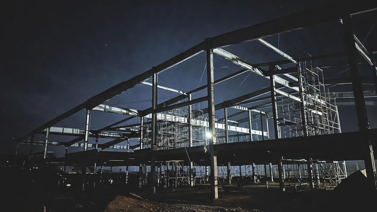 Half-built construction site at night with exposed steel beams and scaffolding illuminated by a single work light from below, casting dramatic upward shadows against a dark sky, empty dirt and gravel ground, no workers present, moody industrial photorealistic style representing an incomplete foundation. Half-built construction site at night with exposed steel beams and scaffolding illuminated by a single work light from below, casting dramatic upward shadows against a dark sky, empty dirt and gravel ground, no workers present, moody industrial photorealistic style representing an incomplete foundation.