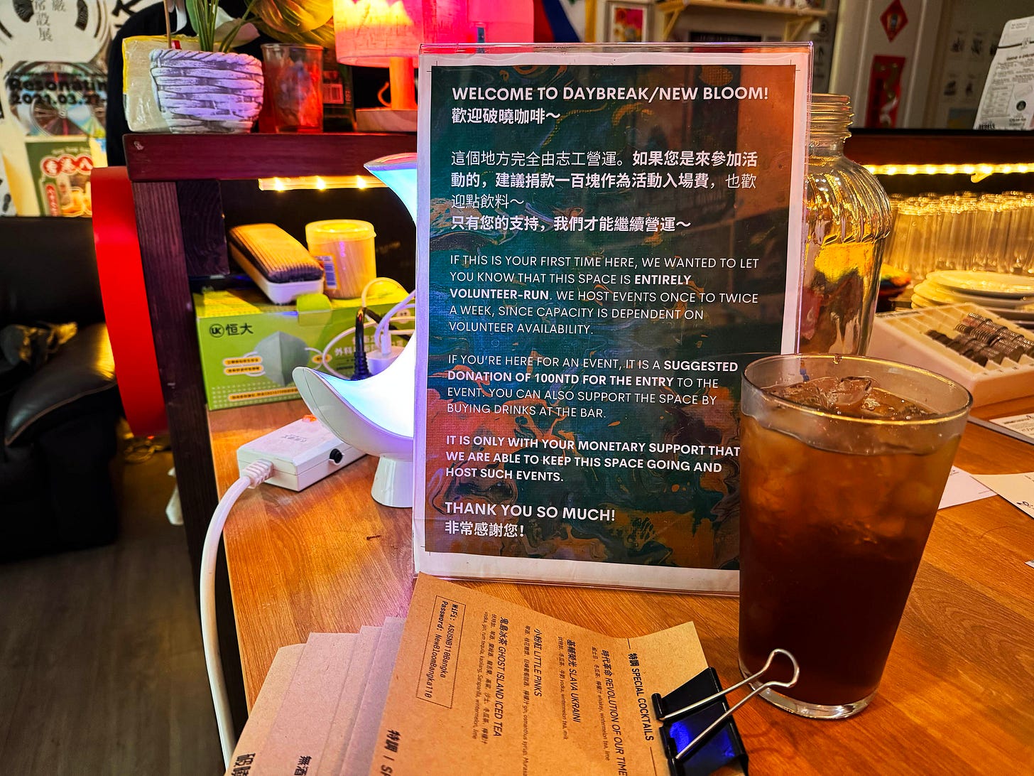 Horizontal photo of the Daybreak bar countertop, with a sign that says Welcome to Daybreak New Bloom and instructions for first-time attendees. The sign is posed next to a small bar menu and a full pint glass. Glassware, a lamp, and a sofa are visible in the background. Horizontal photo of the Daybreak bar countertop, with a sign that says Welcome to Daybreak New Bloom and instructions for first-time attendees. The sign is posed next to a small bar menu and a full pint glass. Glassware, a lamp, and a sofa are visible in the background.