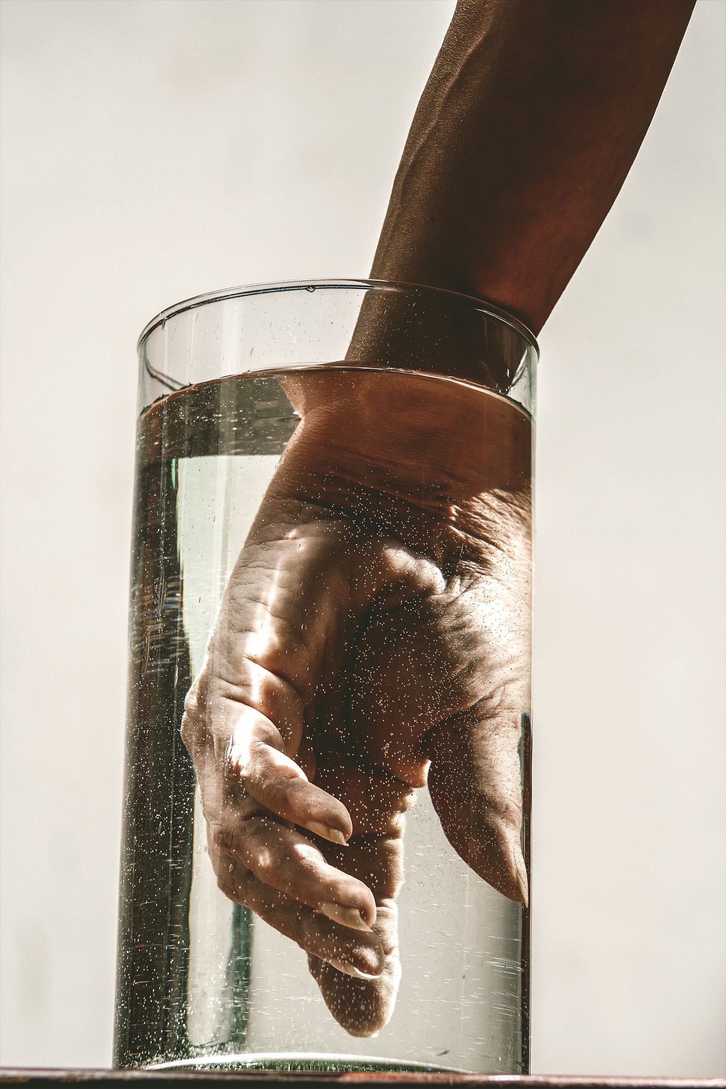 A bare arm submerged in a glass cylinder of water, light catching bubbles along the skin — water as Source, not resource.