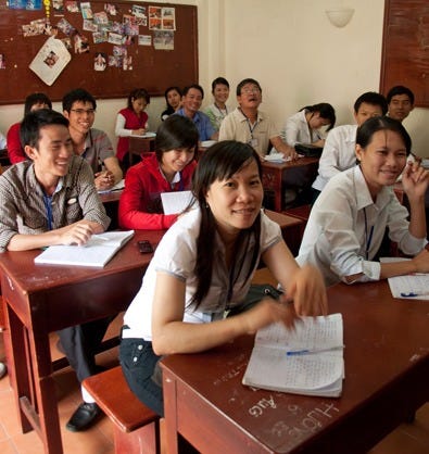 A group of 10 Vietnamese teenagers are sitting at school desks with open notebooks.  