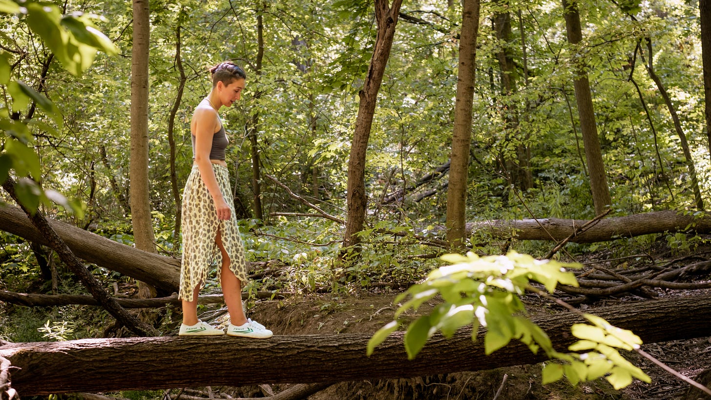 Tarzan in the woods, walking across a long that is suspended four feet above a small creek, her hair braided back and looking down at her feet