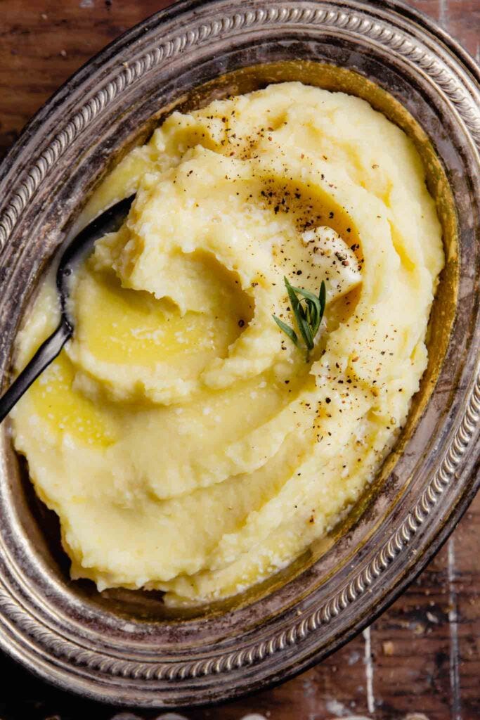 creamy parsnip and potato mash in a silver oval dish with a pad of butter, pepper, salt and tarragon sprinkled over top