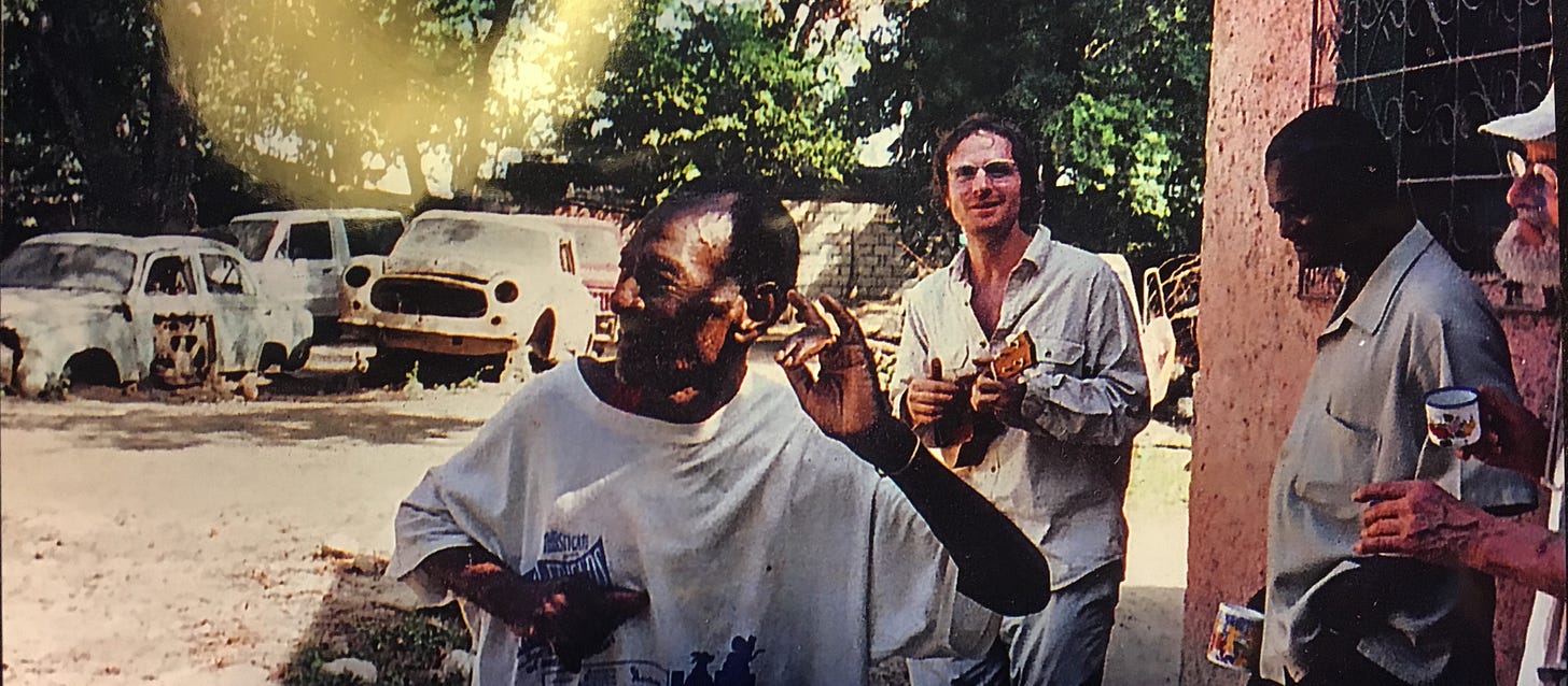 A young Ari Gold plays a ukulele outdoors in Haiti, smiling, while the painter and Vodou priest André Pierre gestures in the foreground. Rusted cars sit under trees behind them, and a bearded man's profile is just visible at the right edge. A young Ari Gold plays a ukulele outdoors in Haiti, smiling, while the painter and Vodou priest André Pierre gestures in the foreground. Rusted cars sit under trees behind them, and a bearded man's profile is just visible at the right edge.