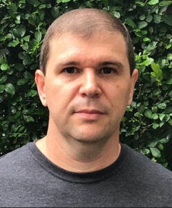 Luciano Freschi - Homem de cabelos pretos e olhos escuros, usando camiseta preta, tendo ao fundo arbustos com folhas verdes