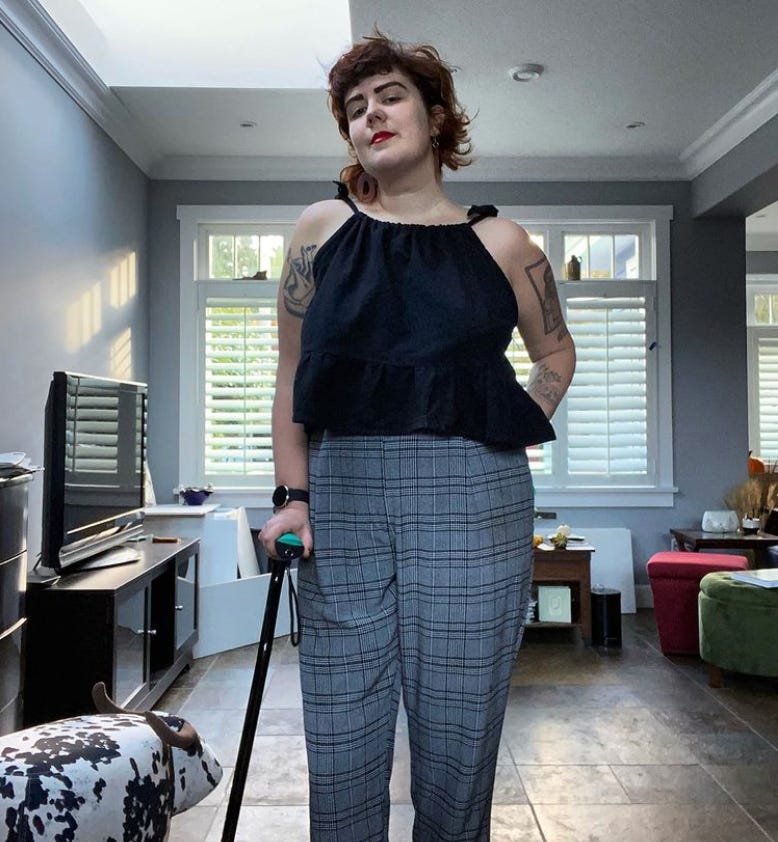 Tash is a tall white woman with brown hair, red lipstick on, and with tattoos on her upper arms and left forearm. She is wearing a black cropped tank top with shoulder ties, and high waisted grey plaid pants. Her left arm is behind her back and her right arm is gripping a black cane with a turquoise handle. She is standing in the middle of a gray room, and there are windows behind her letting a little light through. To her right is a little cow shaped footstool.