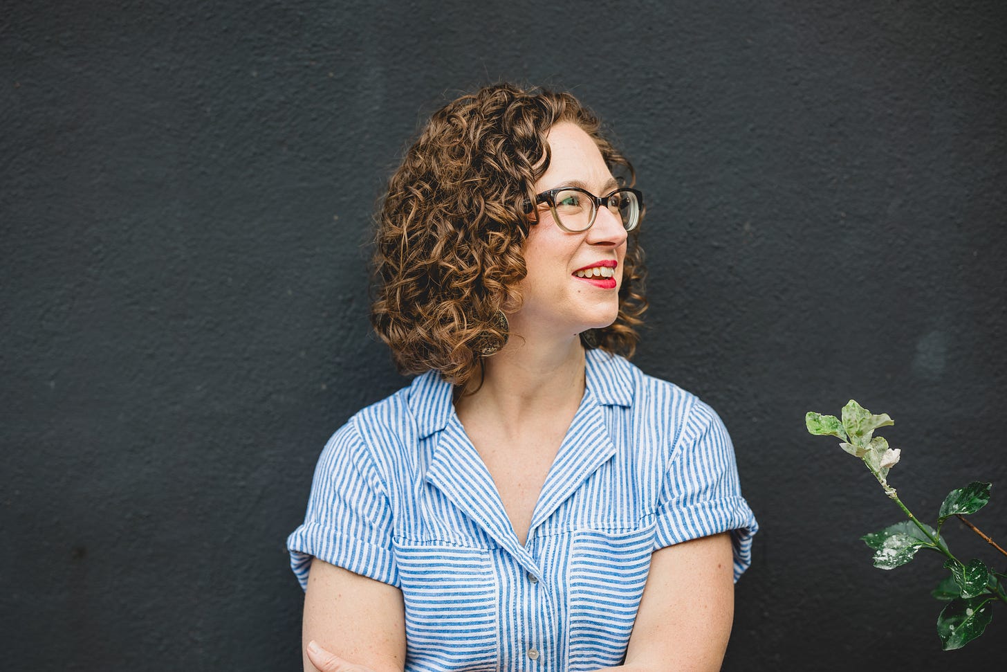 Heather Lanier stands against a dark grey background, gazing off to her left with a slight smile; she wears a blue and white striped short-sleeved shirt, tortoise shell glasses, red lipstick, and her arms crossed; the stem of a plant bends into the photo
