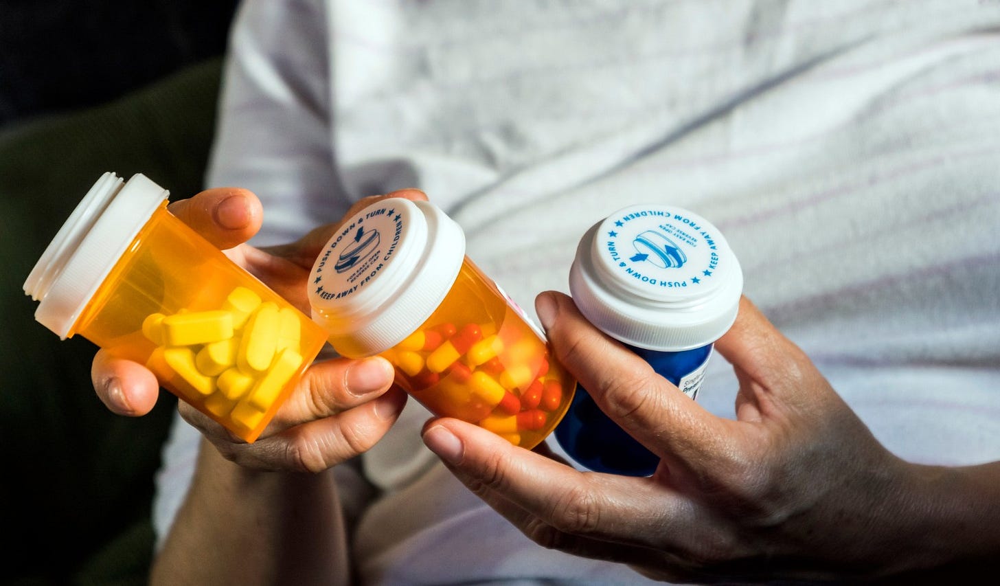 Close-up of a person holding three prescription pill bottles, two orange and one blue, each filled with tablets or capsules. The person’s hands are visible, and the white child-resistant caps feature printed safety instructions. The background is softly blurred, drawing focus to the medication containers.