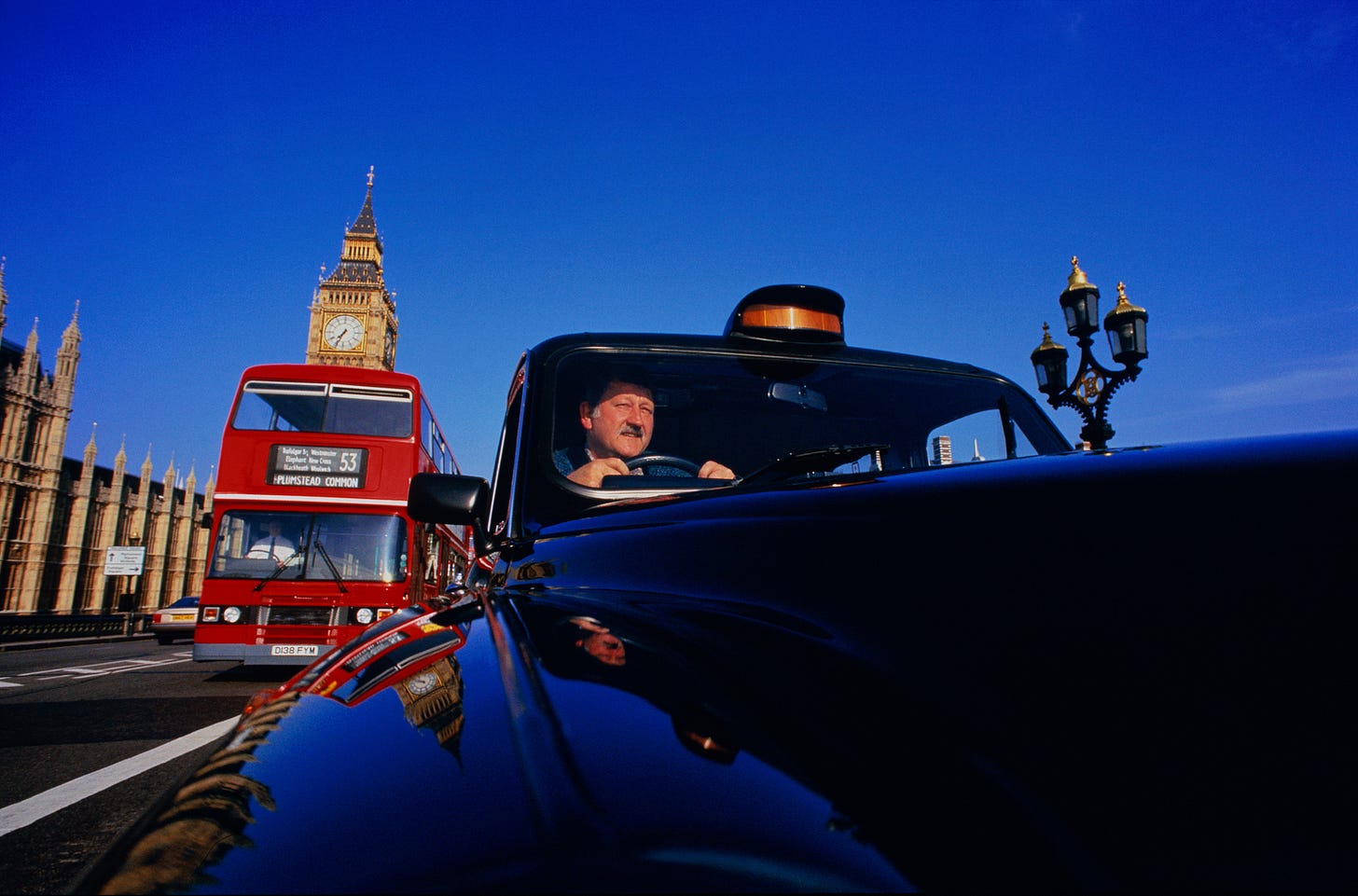 A black cab and a red double-decker bus drive past Big Ben and the Houses of Parliament in London under a clear blue sky. A black cab and a red double-decker bus drive past Big Ben and the Houses of Parliament in London under a clear blue sky.