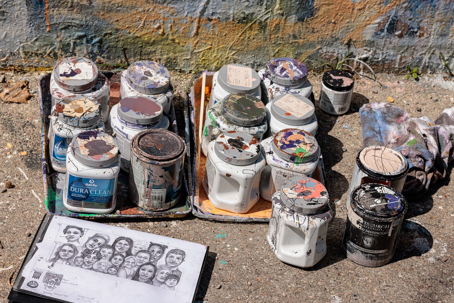 A collection of paint-stained jars and cans arranged on cardboard, next to an open sketchbook with a pencil drawing of the mural's row of faces.