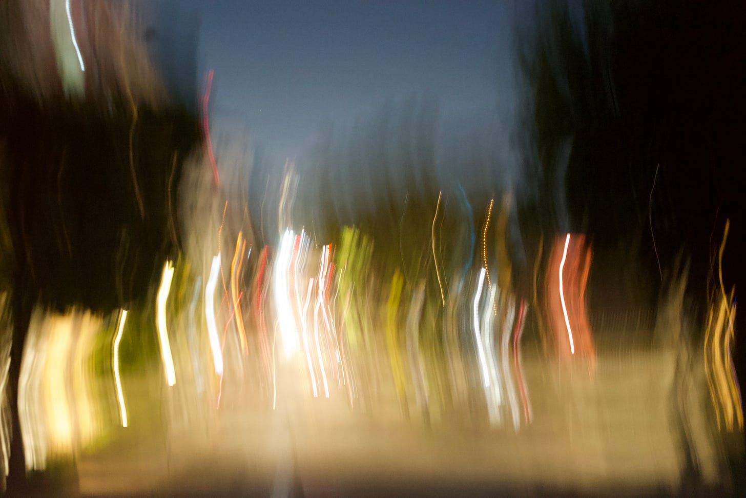Long exposure photograph of a nighttime street scene rendered in soft motion blur, with streaks of white, red, and amber lights stretching vertically through the frame. The abstract composition evokes movement, disorientation, and the feeling of a city in motion.